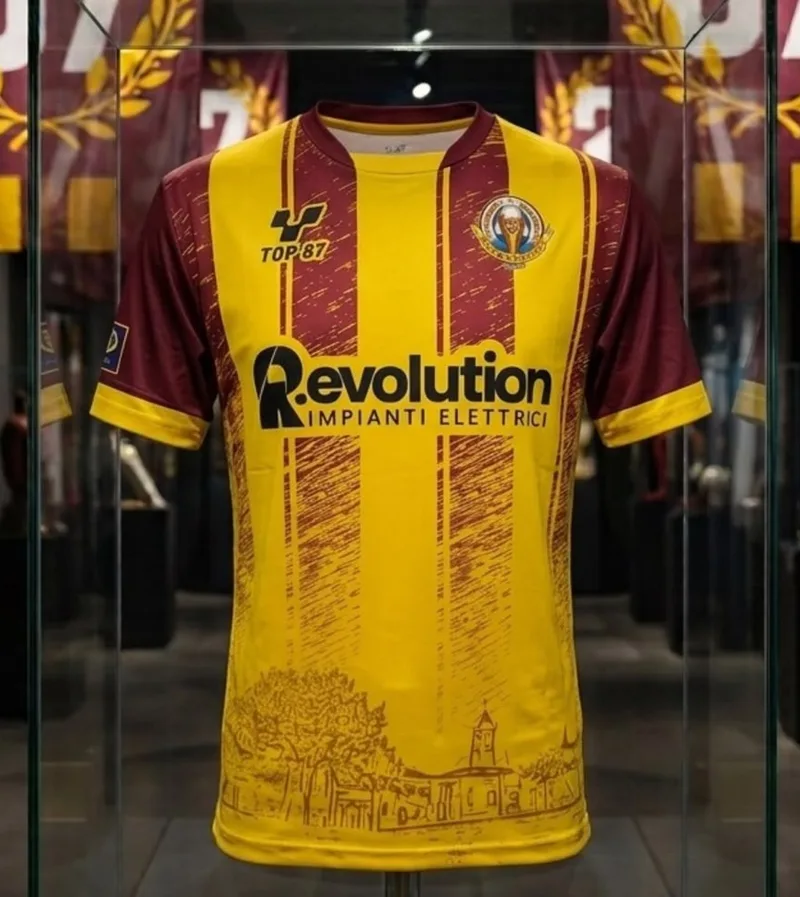 Maglia gialla con bande granata, lo sponsor Revolution e il disegno della piazza del paese in basso.